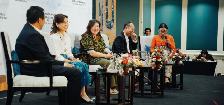 A group of panelists is sitting on a stage, engaging in a lively discussion during the Bali Beyond and Travel Fair (BBTF) 2024. The backdrop displays names and titles of the speakers, and there are floral arrangements on the tables in front of them. The panelists appear to be in high spirits, sharing smiles and laughter.